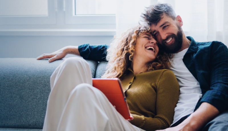 A couple sitting together on a sofa in a living room, smiling at each other.